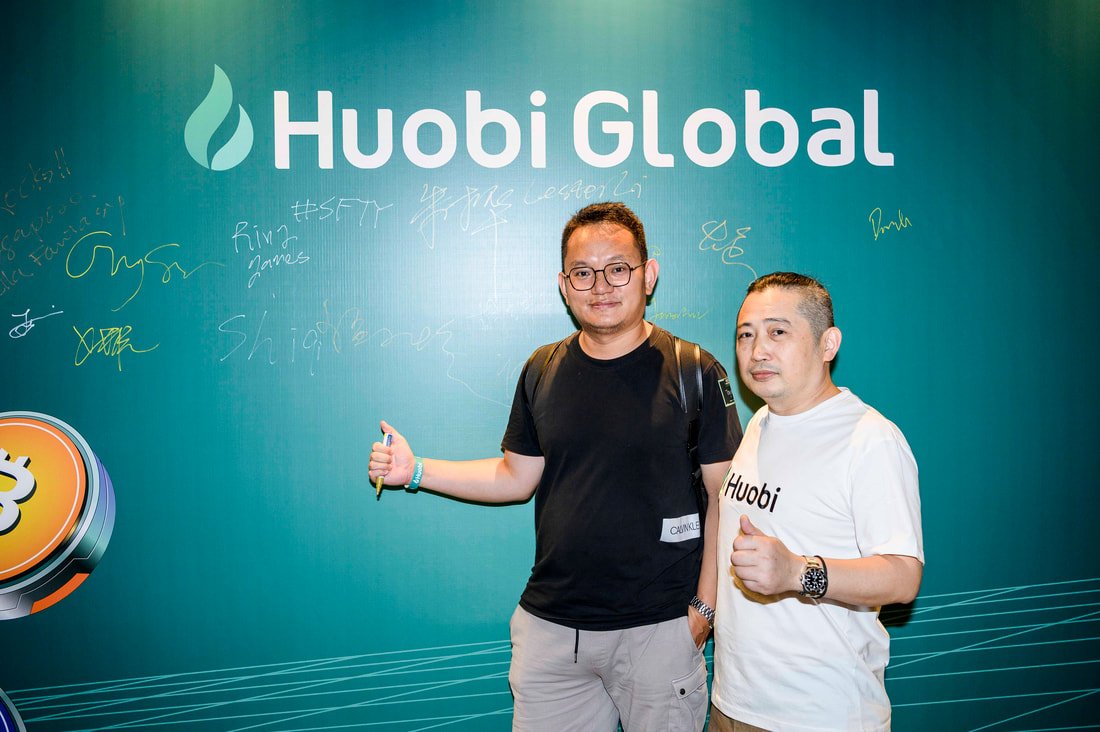 Two men standing in front of a teal Huobi Global banner, both giving a thumbs-up. One is wearing glasses and a black t-shirt, the other is wearing a white Huobi t-shirt. The banner features signatures or messages in various colors.