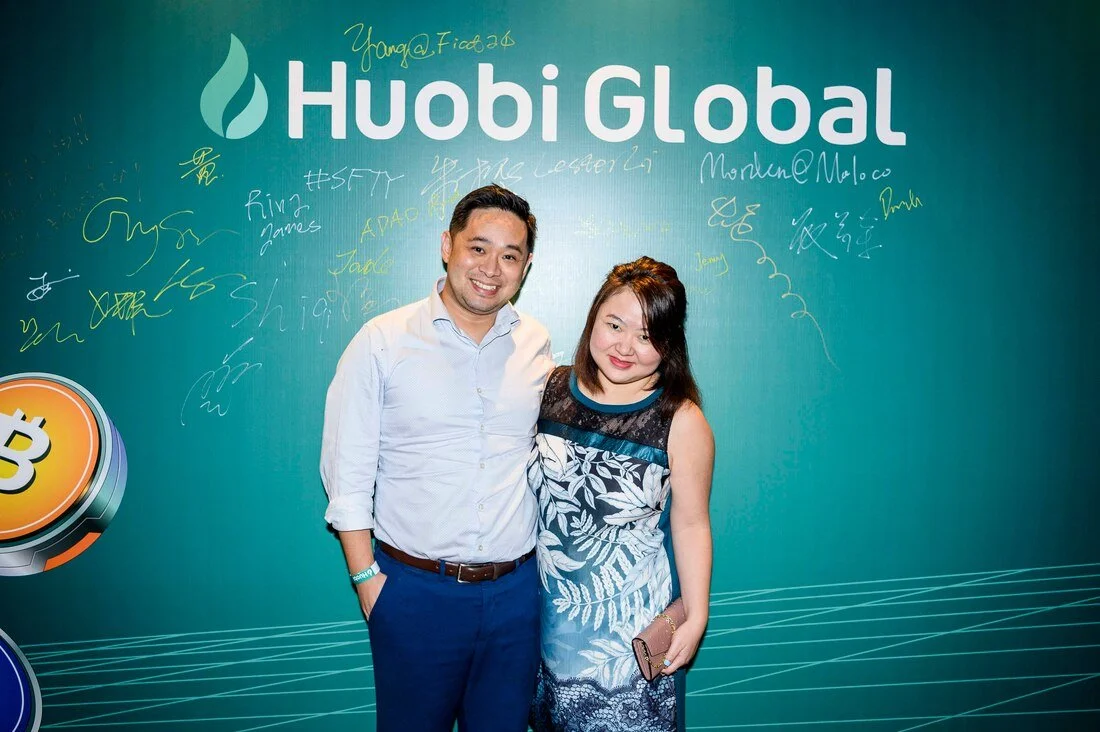 A man and woman standing together in front of a turquoise backdrop with 'Huobi Global' logo, signatures, and signatures on it.