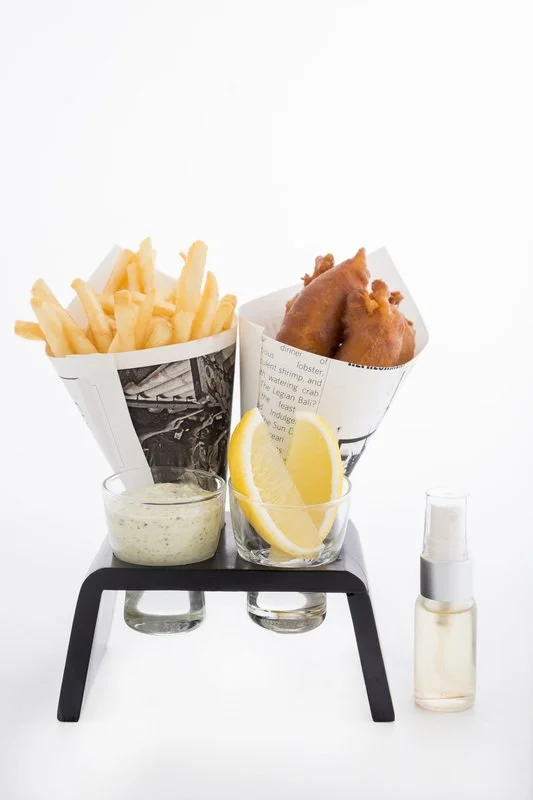 French fries, fried fish fillet, lemon slices, tartar sauce, and a small spray bottle on a black stand against a white background.