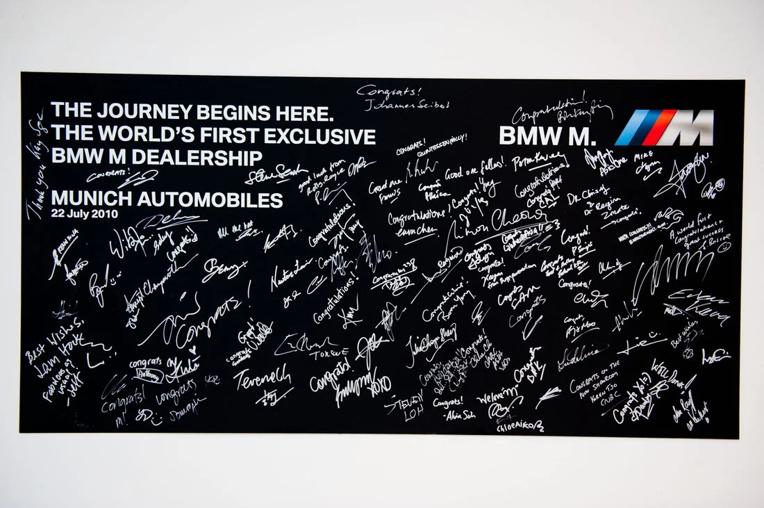 Black signed banner for BMW M dealership opening at Munich Automobiles on July 22, 2010, with numerous handwritten congratulatory notes and signatures.