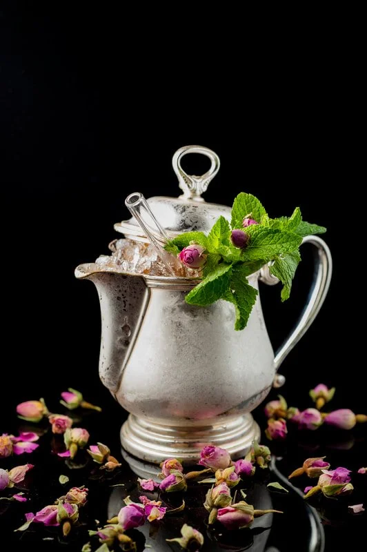 A silver teapot with a lid, garnished with green mint leaves and purple flowers, surrounded by scattered pink flower buds on a reflective black surface against a black background.