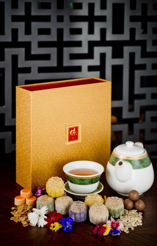 Tea set with teapot, teacup, assorted mooncakes, and colorful flowers on a table with decorative background and gold box