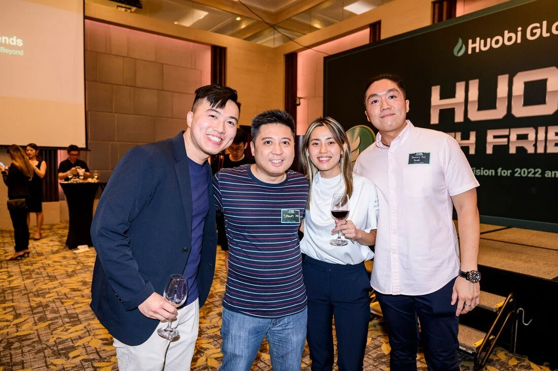 Four people smiling, standing together at a corporate event, holding wine glasses, with a banner that reads 'Huobi Global' in the background.