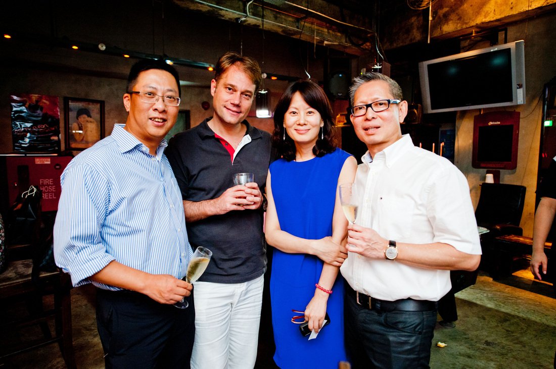 Four people standing together at a social event, holding drinks, smiling for the camera in a dimly lit indoor setting.