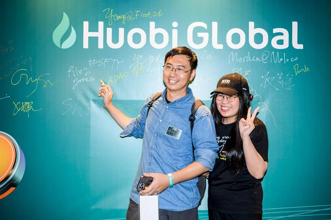 Two people, a man and a woman, smiling and posing in front of a teal wall with 'Huobi Global' text and signatures. The man is wearing glasses, a blue shirt, and has a backpack, while the woman is wearing glasses, a black cap, and a black t-shirt. The