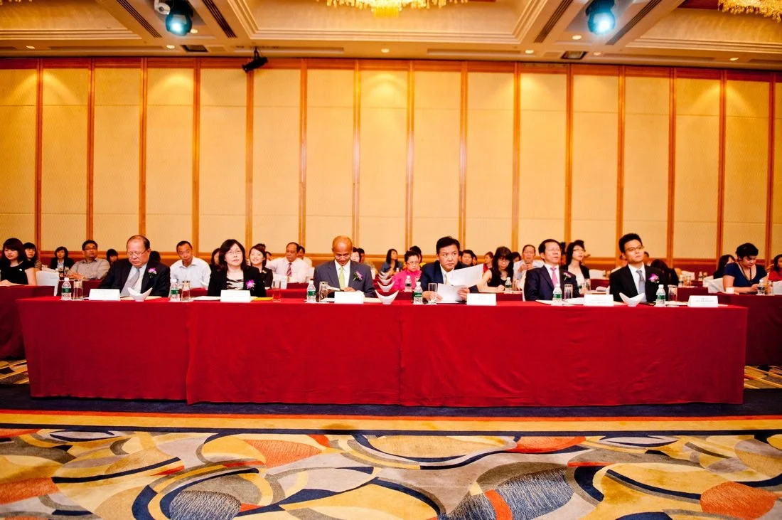 Conference or meeting with people sitting at a long table covered with a red tablecloth in a large room with a patterned carpet.
