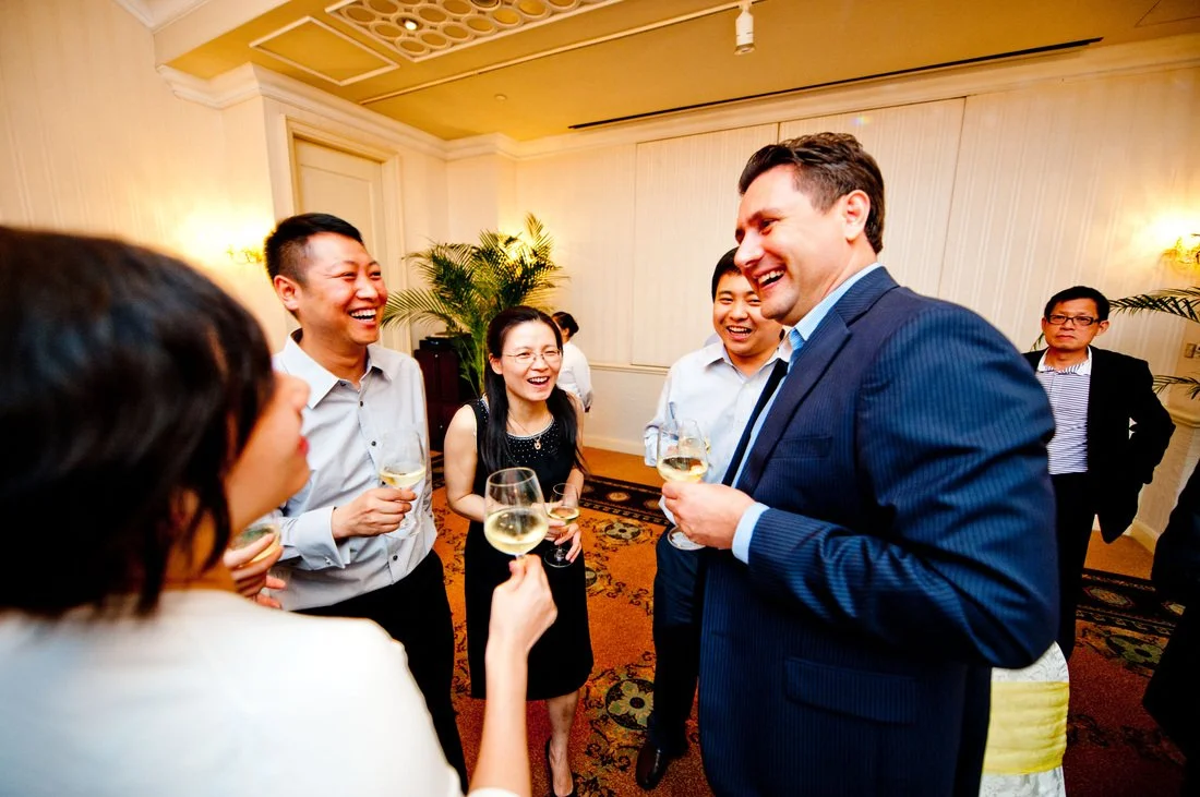 Group of six people in formal attire, laughing and chatting at a social gathering, holding wine glasses in a decorated room with plants and warm lighting.