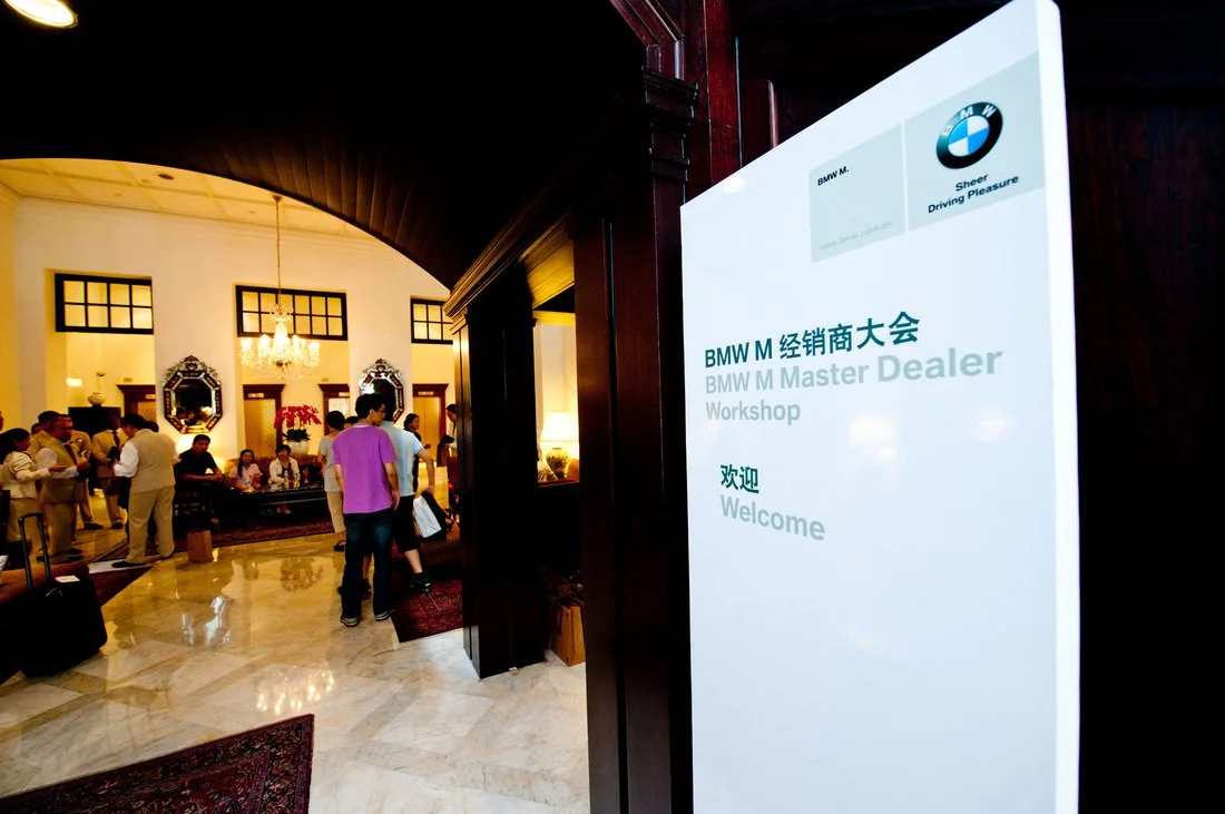 Hotel lobby with a large chandelier, ornate mirrors, and guests checking in or waiting. A sign reads 'BMW M Master Dealer Workshop Welcome' in Chinese and English.