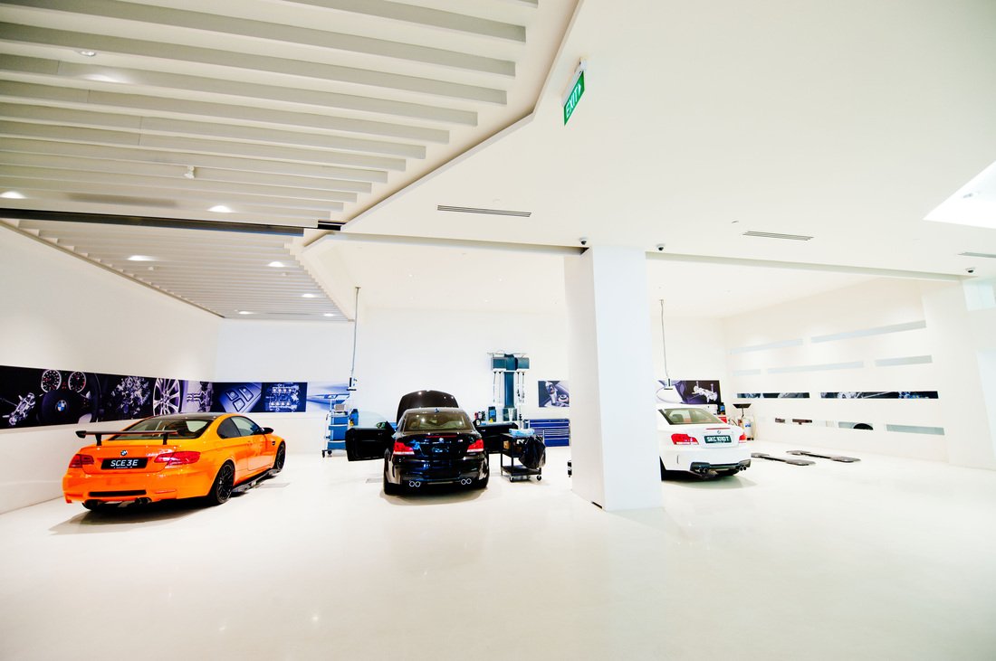 A spacious, white-walled garage with three sports cars: an orange car on the left, a black car in the center, and a white car on the right, with tools and equipment nearby.