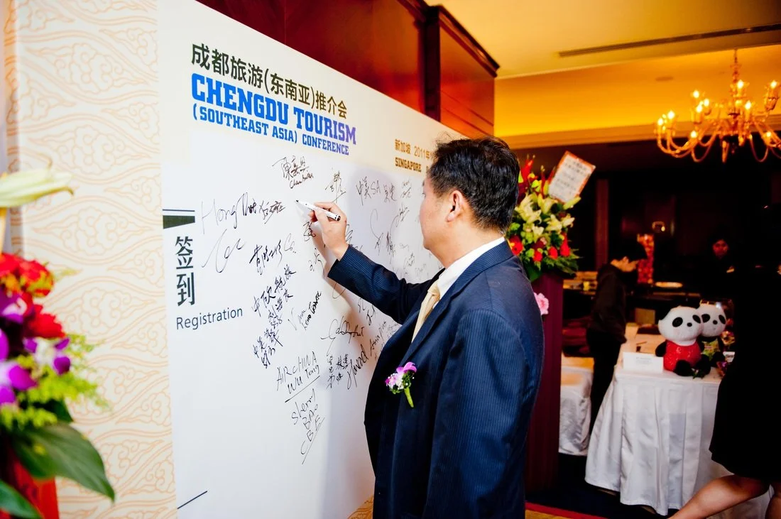 Man in a blue suit signing on a large white board at the Chengdu Tourism Southeast Asia Conference, with floral arrangements and panda plush toys in the background.