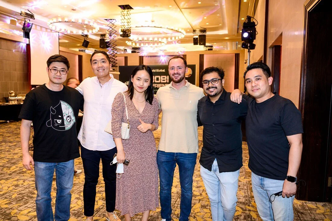 Group of seven people smiling at a social event, standing on a patterned carpet in a banquet hall with chandeliers and stage lights.