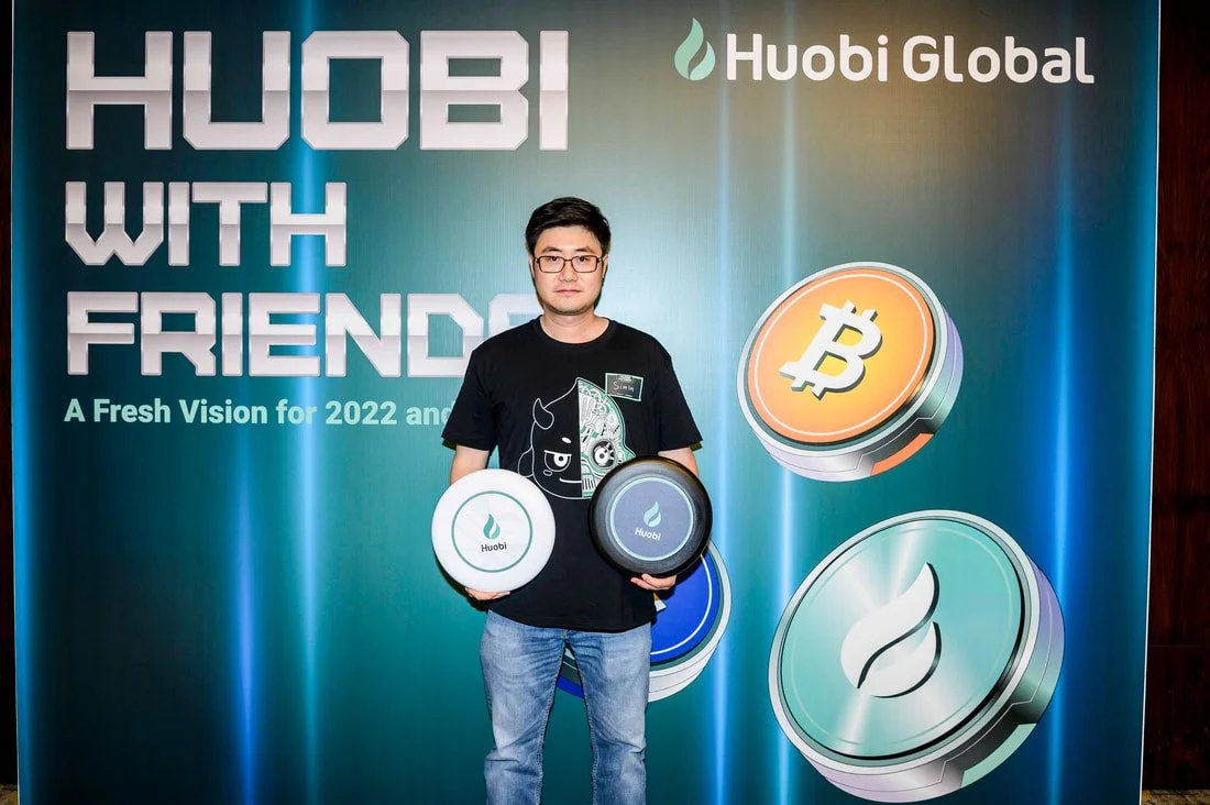 Man standing in front of a Huobi Global banner holding cryptocurrency coins, with Bitcoin and other cryptocurrency logos displayed.