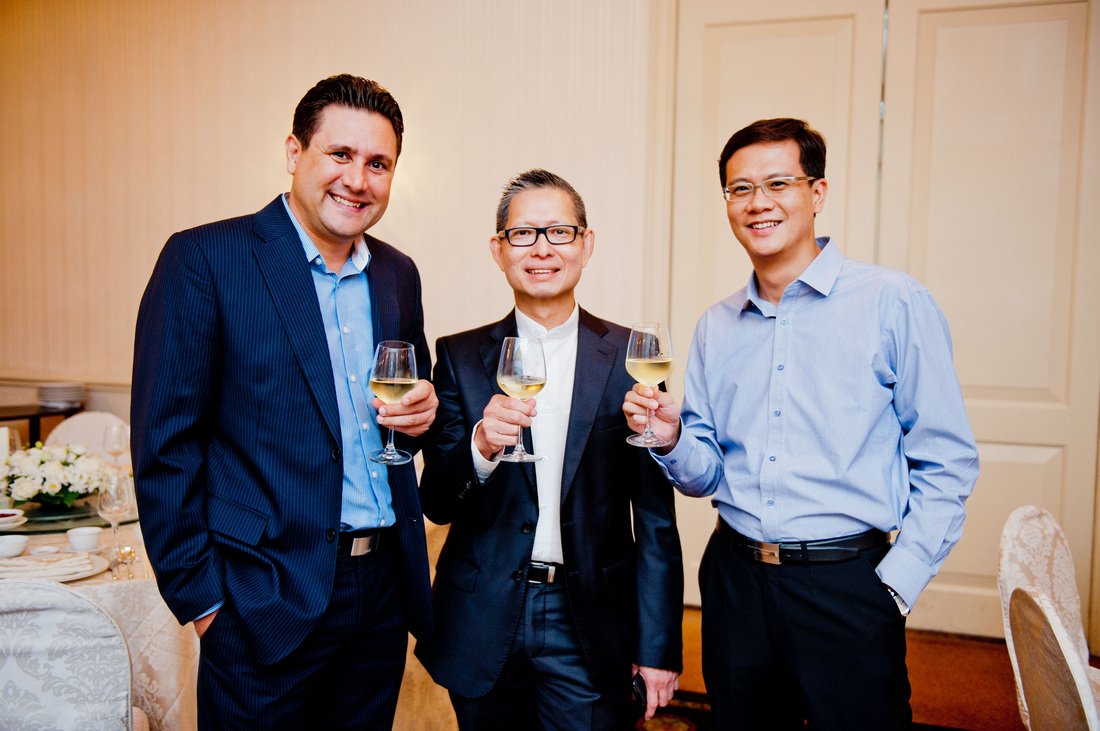 Three men in suits holding glasses of white wine at a formal event.