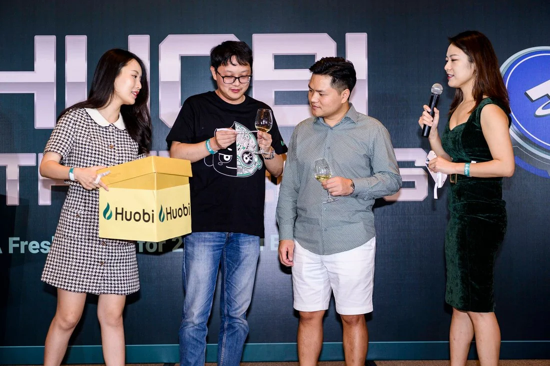 Four people on a stage, two women and two men, at a corporate event. One woman holds a yellow box labeled 'Huobi,' and the others hold wine glasses. A woman on the right speaks into a microphone.
