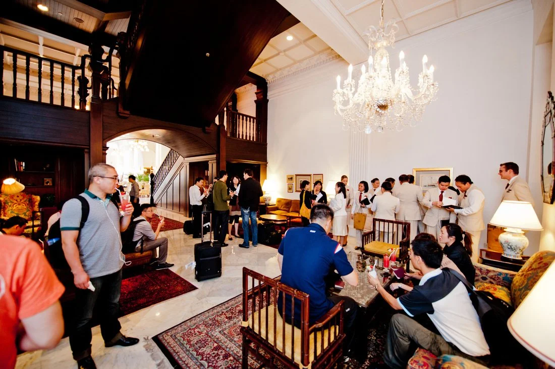 Hotel lobby with a chandelier, wooden staircase, and people waiting and socializing, some with luggage.