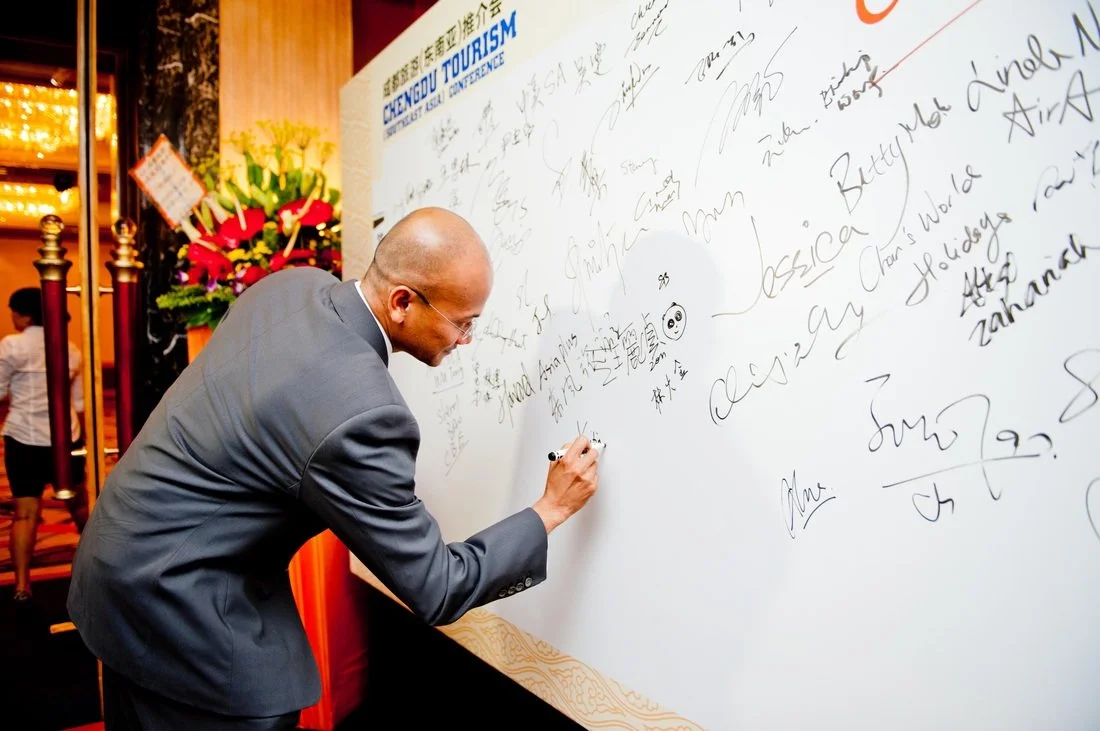 A man in a gray suit signing a large white board filled with signatures and messages at an event.