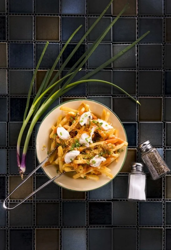 A bowl of pasta with white sauce, topped with sour cream, chopped green onions, and herbs, placed on a dark tiled surface with a bunch of green onions and two seasoning shakers nearby.