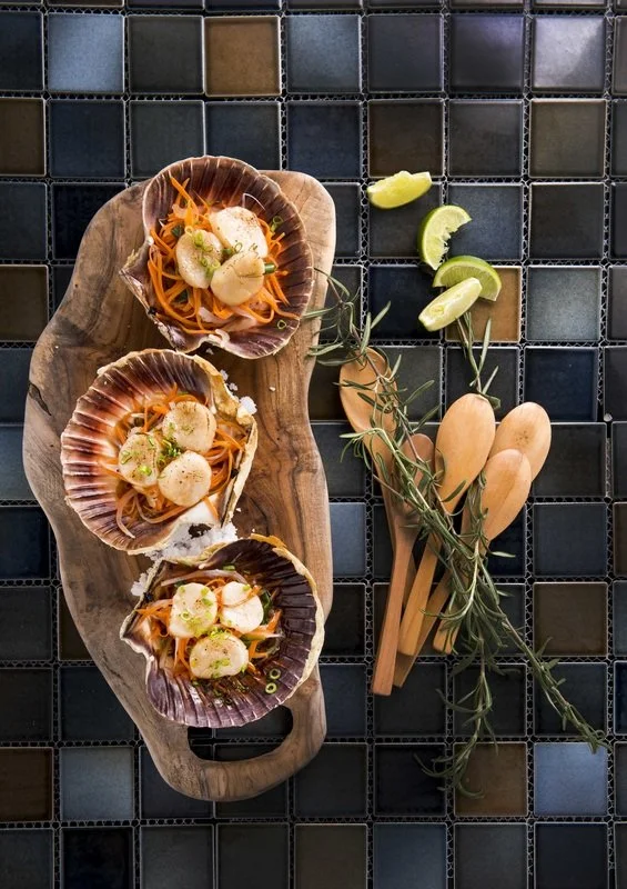 Three scallop dishes served on shells placed on a wooden serving board, with lime wedges, wooden spoons, and sprigs of herbs on a tiled surface.