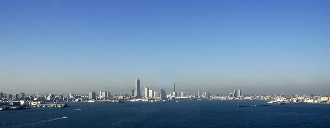 Panoramic view of a city skyline with tall modern buildings across a large body of water under a clear blue sky.