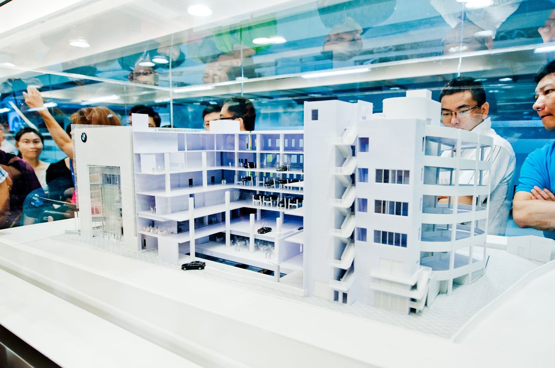 Architectural model of a modern multi-story building displayed behind glass at a public exhibition, with visitors observing.