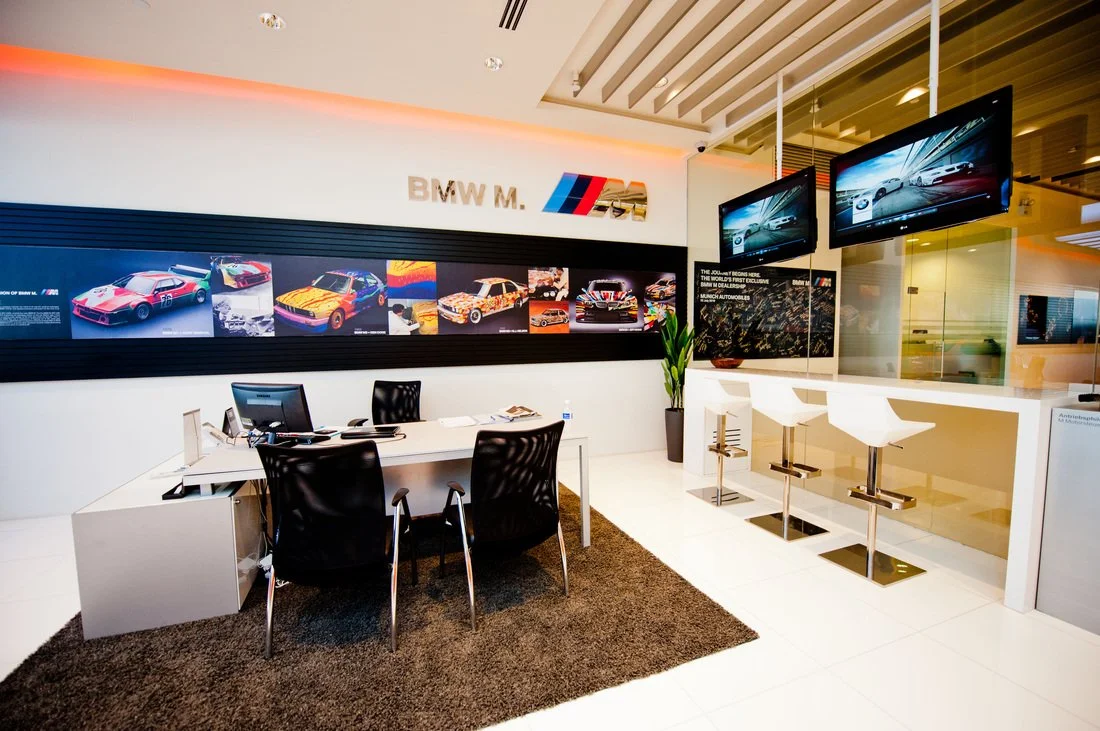 BMW dealership interior with a reception desk, black chairs, two mounted screens showing BMW cars, and BMW M series branding on the wall.