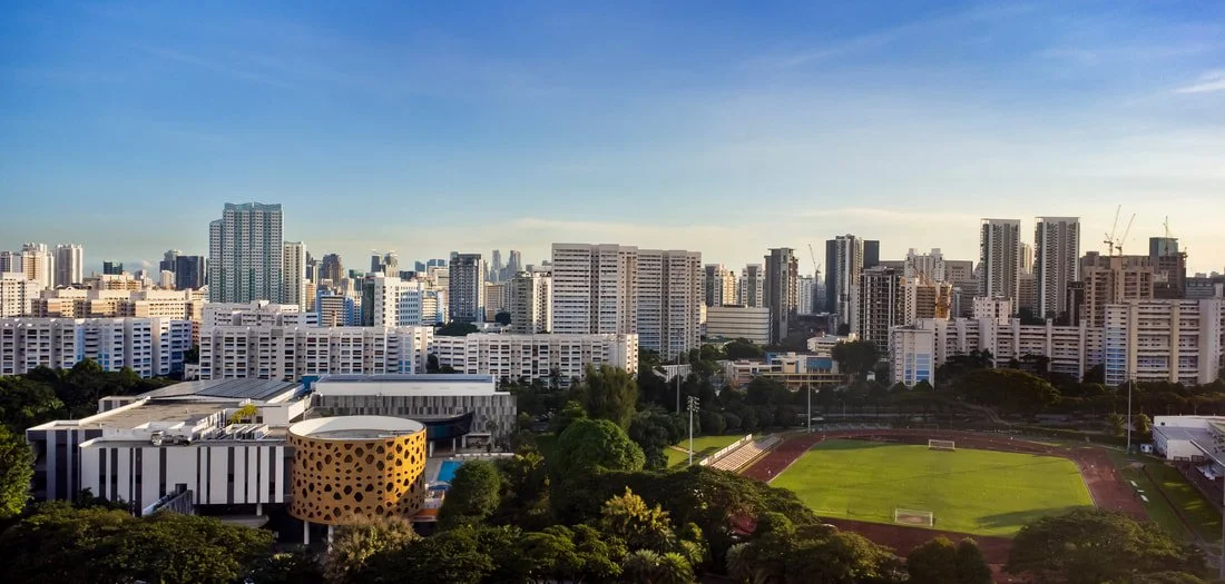 City skyline with numerous high-rise buildings, a sports field, and greenery, under a blue sky in the late afternoon.