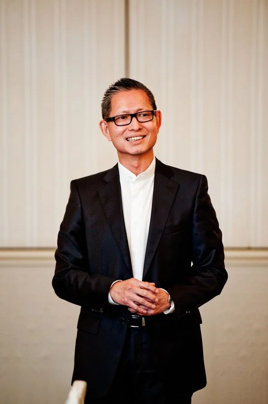A man wearing a black suit, white shirt, and glasses standing in front of a beige curtain, smiling with his hands clasped.