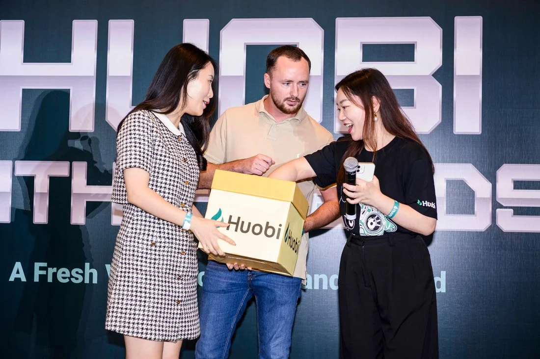 Three people on stage at an event, two women and one man, with a Huobi box. The woman on the right is smiling and holding a microphone with her arm extended into the box, while the woman on the left helps hold the box. The man watches with a serious 