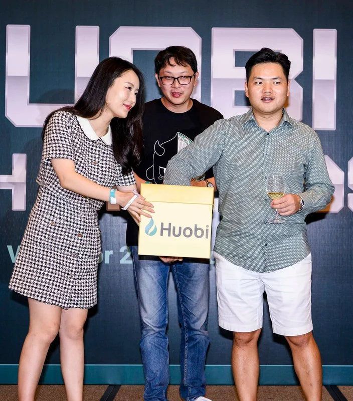 Three people at a Huobi event, with two men and a woman, standing in front of a dark blue background with large white letters. The woman is holding a huobi box, and the man on the right is holding a glass of white wine.