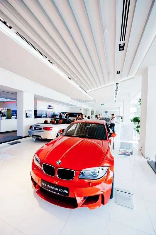 Red BMW M Series coupe on display in a modern car showroom with other vehicles and people in the background.