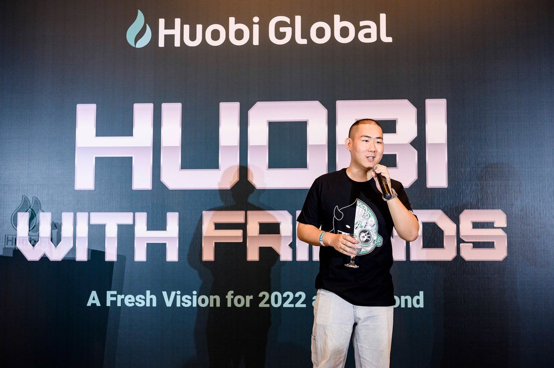 A man in a black T-shirt holding a microphone and a glass of water, standing in front of a Huobi Global event backdrop with the text "HUB! WITH FRIENDS" and a slogan about a fresh vision for 2022.