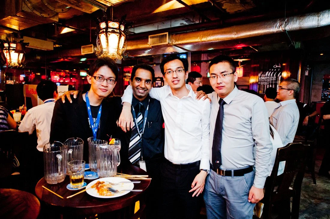 Four men in formal attire posing together at a restaurant or bar, with dishes and drinks on the table in front of them, and other people in the background.