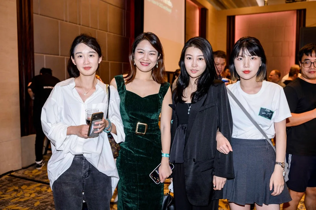 Four young women standing together at an event, dressed in stylish casual and formal attire, with a background of a patterned carpet and other people attending the event.