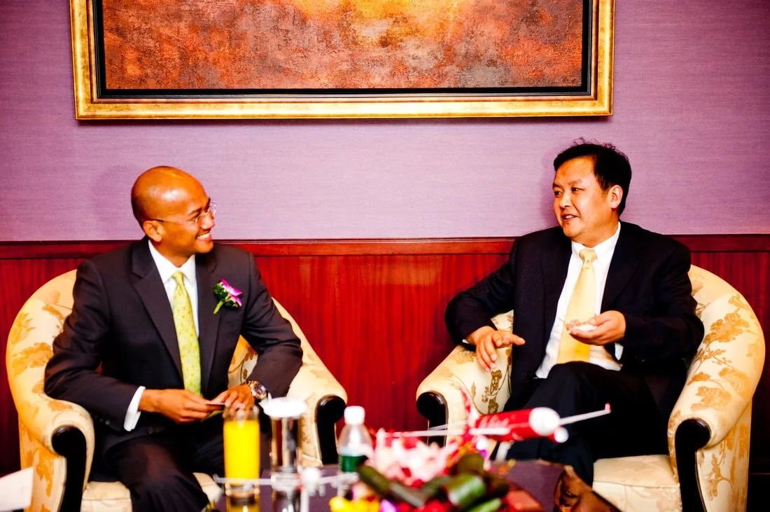 Two men in suits having a conversation in a sitting room with purple walls and a framed painting above them. The man on the left is smiling, holding a small object, and wearing glasses, while the man on the right is speaking and gesturing with his ha