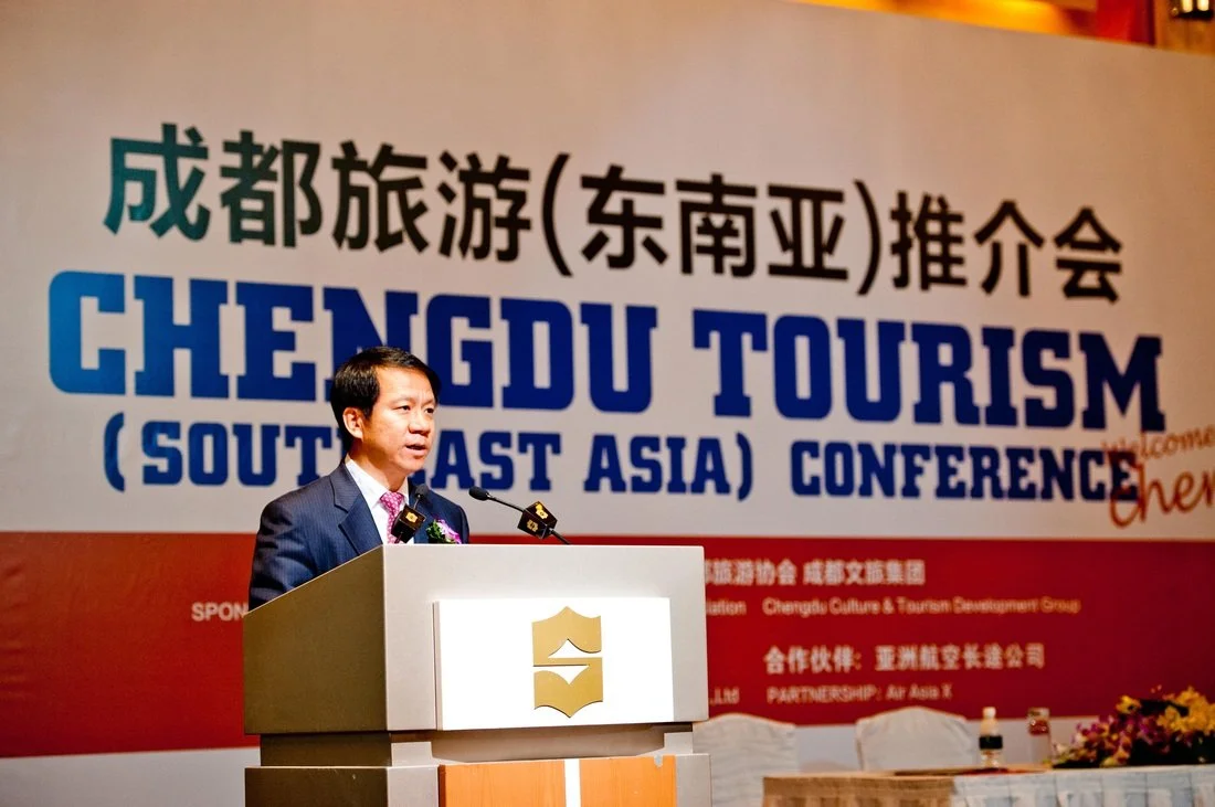 Man in a suit speaking at a podium during a conference about Chengdu tourism, Southeast Asia conference, with a large event banner in the background.