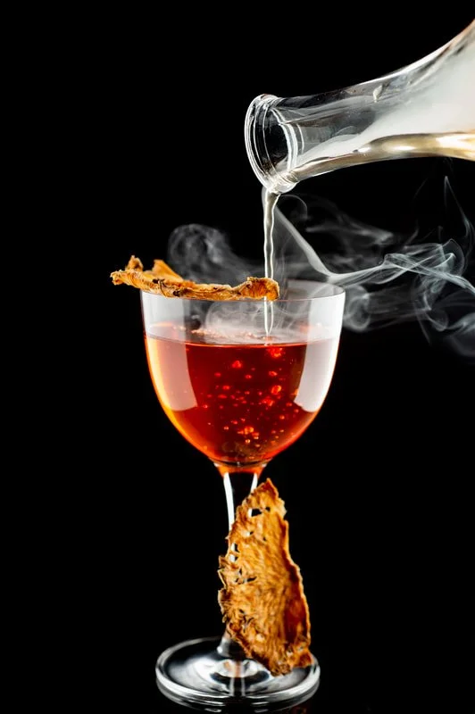 A glass of red wine with a piece of dried fruit on the rim, being poured with clear liquid from a tilted glass pitcher, with a black background.