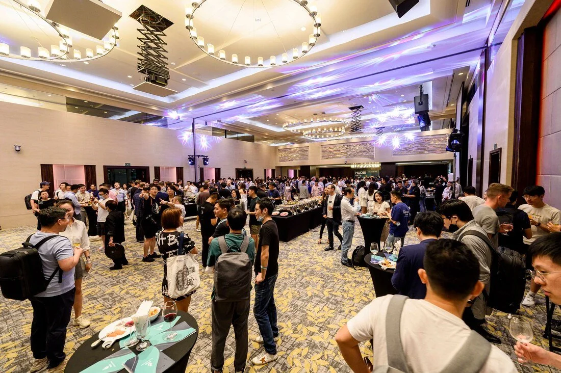 Crowd of people at a large indoor event or conference, standing around tables with drinks and food, in a well-lit hall with decorative ceiling lights and purple lighting effects.