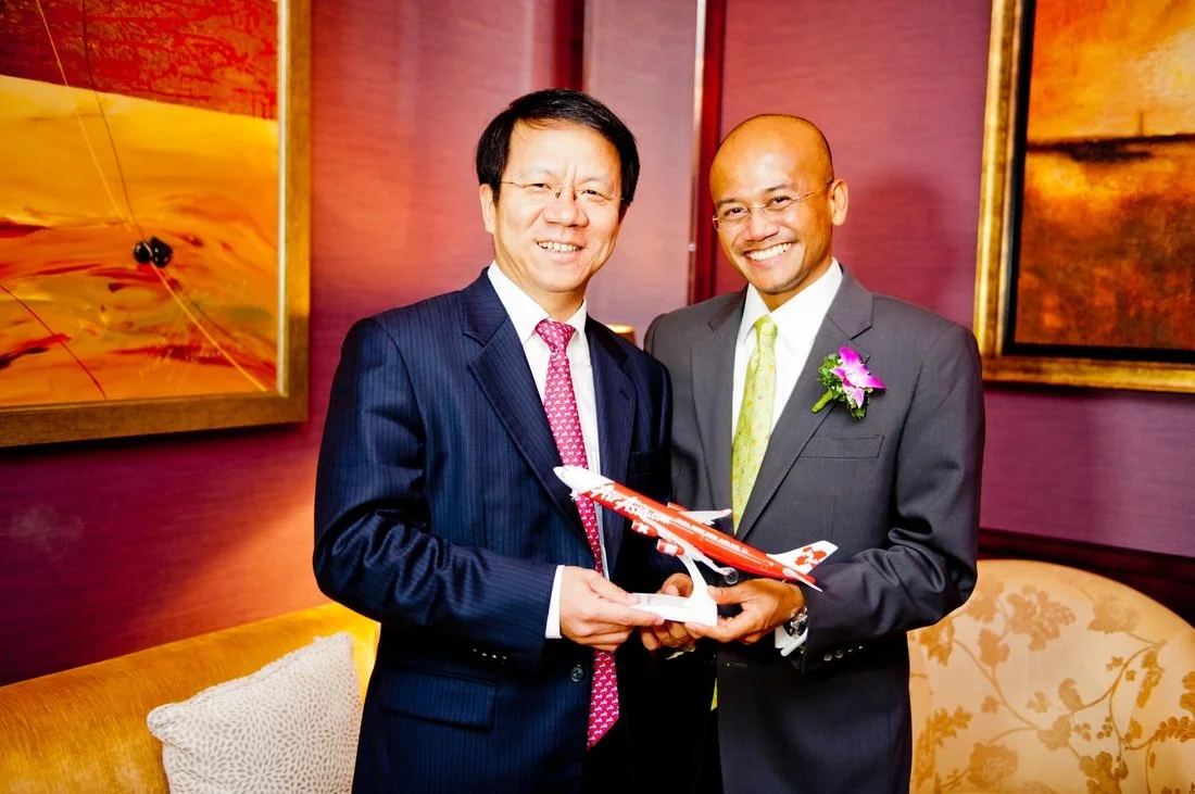 Two men in suits smiling and holding a model airplane together at an indoor event.