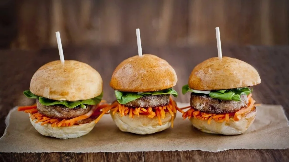 Three mini hamburgers with seasoned beef patties, shredded carrots, lettuce, and soft buns arranged on parchment paper on a wooden surface.