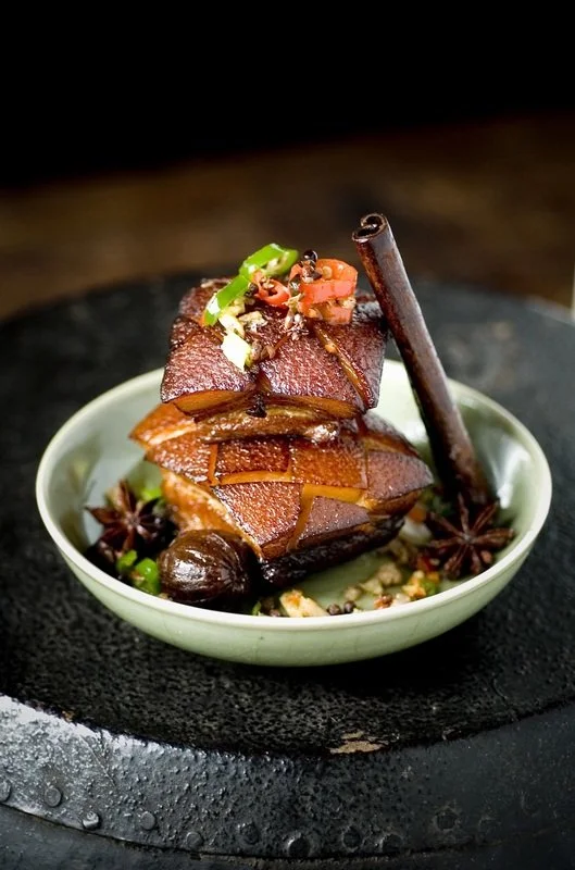 Close-up of braised pork belly garnished with green onion, star anise, and a cinnamon stick in a small bowl on a black textured surface.