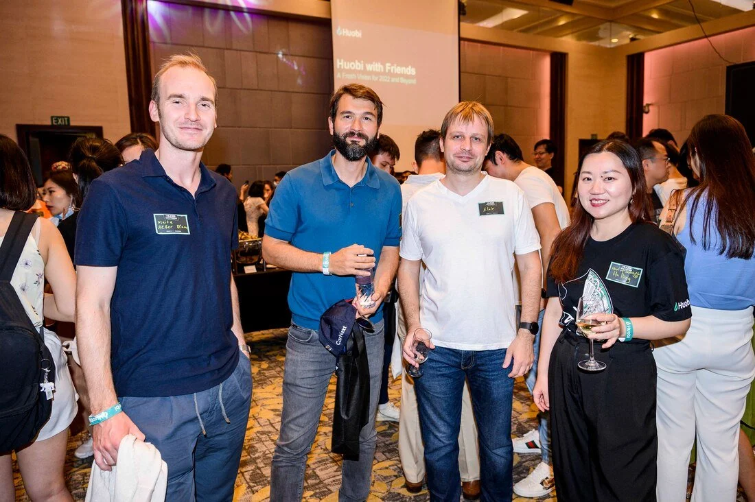 Four people standing together at a conference or social event, holding drinks, with a crowd in the background and a presentation screen behind them.