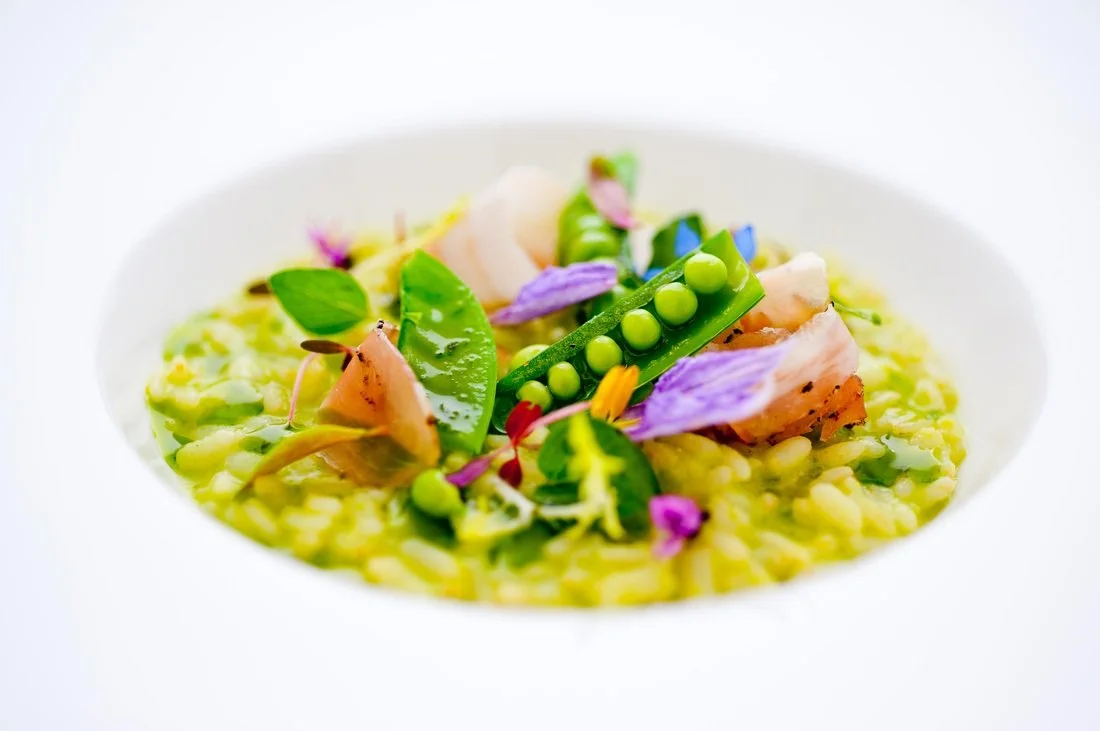 A white bowl filled with gourmet risotto topped with peas, edible flowers, snap peas, and small pieces of cooked meat.