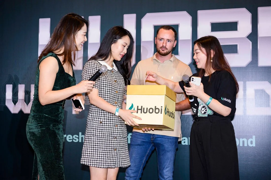 Four people on stage, three women and one man, participating in a presentation or event. One woman in black dress is handing a yellow box with the Huobi logo to a woman in a black and white checked dress. The man and another woman are watching, with 