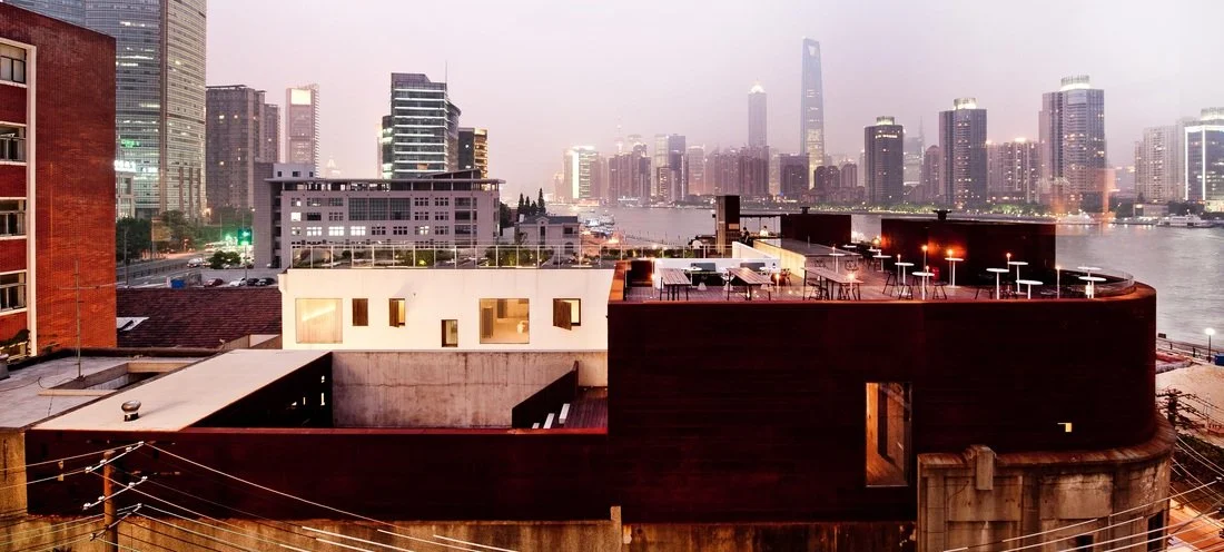 City skyline at dusk with high-rise buildings along a river, some lit, with a rooftop terrace in the foreground.