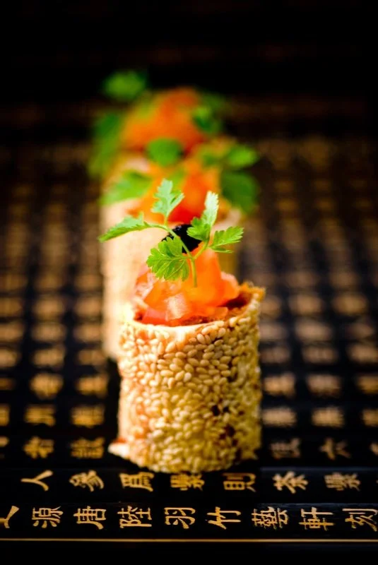 Close-up of a sushi roll topped with salmon, cilantro, and green garnishes, with a background of black and gold patterned fabric featuring Chinese characters.