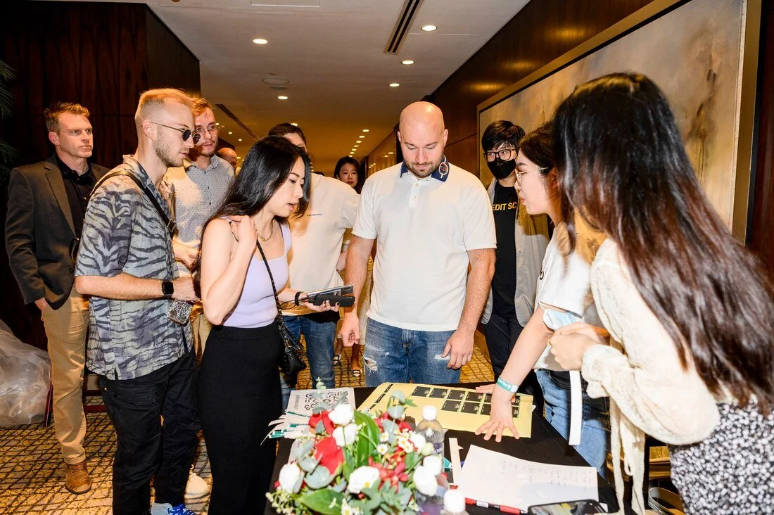 Group of people gathered around a table at an indoor event, looking at items and notes displayed on the table.