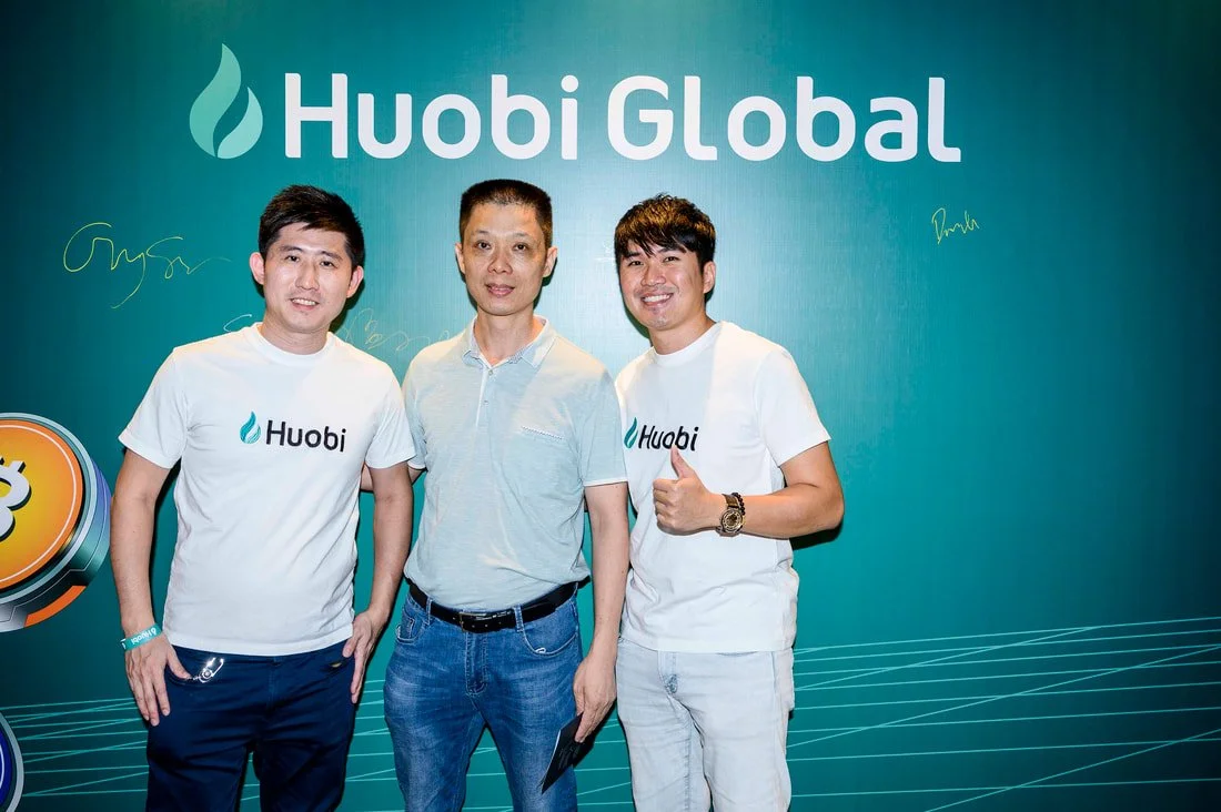 Three men standing in front of a teal background with the words "Huobi Global" and its logo, posing for a photo at a company event.