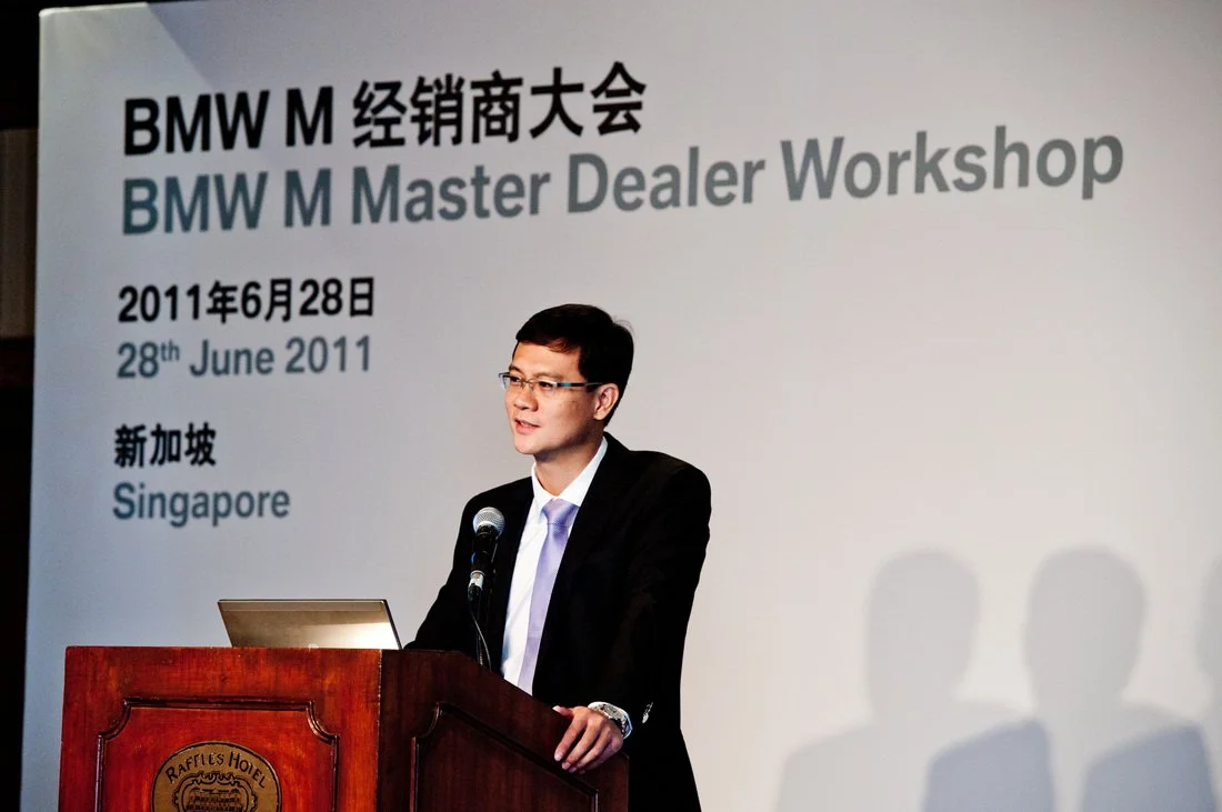 A man in a suit and glasses is speaking at a wooden podium with a microphone during a BMW M Master Dealer Workshop on June 28, 2011, in Singapore, with a large presentation screen behind him displaying event details.