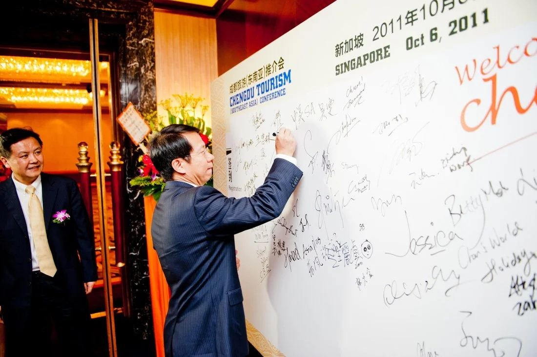 Man in a suit signing a large guestboard at the Chengdu Tourism conference in Singapore, October 6, 2011, with another man watching. The guestboard is filled with signatures and messages.