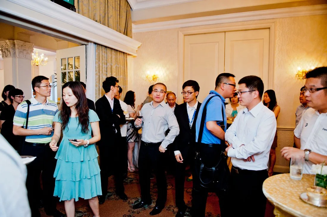 Group of people in formal attire socializing at an indoor event or reception, with some standing in small groups, holding drinks, in a well-lit room with cream-colored walls and decorative lighting.